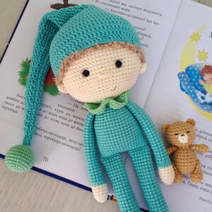 May include: A crocheted doll with a teal blue body and a matching hat. The doll has a white face with brown hair and black eyes. The doll is holding a small brown crocheted teddy bear. The doll is sitting on a book with text in a foreign language.