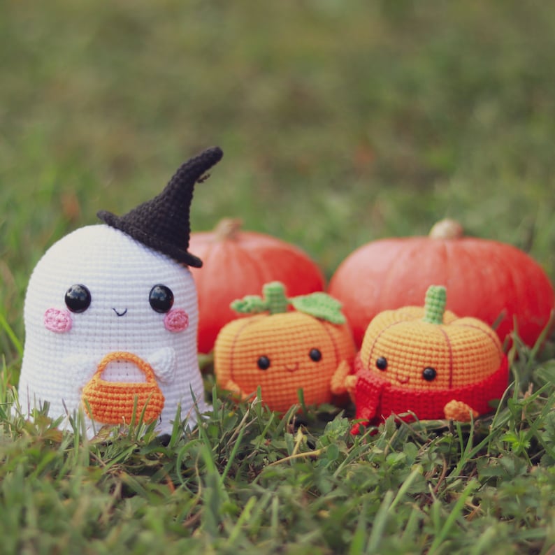 May include: A white crocheted ghost wearing a black witch hat holds a small orange crocheted pumpkin. Three orange crocheted pumpkins with smiling faces sit in the grass.