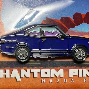 May include: A blue enamel pin of a vintage Mazda RX3 car with a desert landscape background. The pin reads "PHANTOM PINS MAZDA RX3".