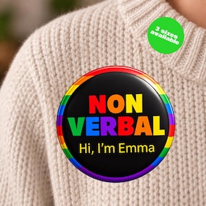 May include: A round pinback button with a black background and a rainbow-colored border. The text reads "NON VERBAL" in large, multi-colored capital letters, followed by "Hi, I'm Emma" in yellow text. A green circle indicates 3 sizes available.