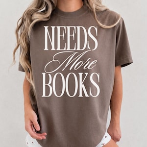 Puede incluir: Una camiseta marrón de manga corta con el texto blanco "NEEDS More BOOKS" impreso en una combinación de fuentes serif en negrita y cursiva elegante en el pecho. La prenda tiene un cuello redondo y una silueta holgada y relajada.