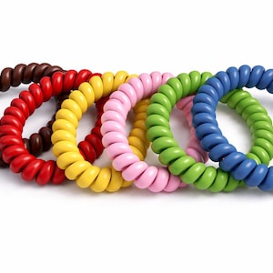 Könnte beinhalten: Eine Sammlung von sechs spiralförmigen Haargummis in verschiedenen Farben: Braun, Rot, Gelb, Rosa, Grün und Blau. Diese farbenfrohen, spiralförmigen Haaraccessoires sind auf weißem Hintergrund angeordnet.