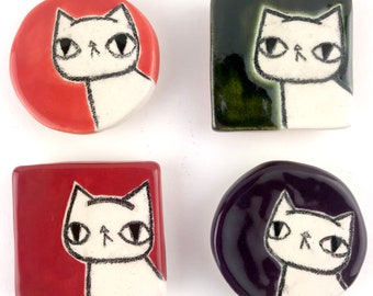 May include: Four ceramic cat magnets in different colors: red, green, purple, and orange. Each magnet features a white cat with black eyes and a black nose.