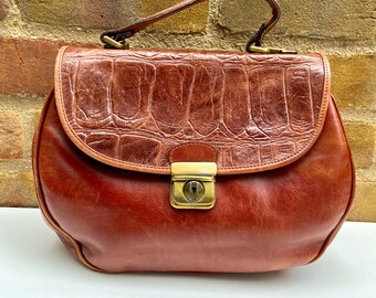 May include: A brown leather handbag with a top flap and a gold clasp. The bag has a textured crocodile-embossed design.