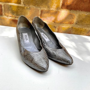 Può includere: Un paio di scarpe con tacco in pelle di serpente grigia con punta a punta. Le scarpe hanno un logo Bally all'interno di ogni scarpa.