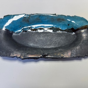Pode incluir: Um prato de cerâmica feito à mão com formato retangular e interior oval raso. A borda superior é esmaltada em um turquesa vibrante, contrastando com o interior cinza escuro. O prato tem um toque rústico e orgânico.
