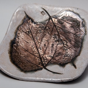 Puede incluir: Un plato de cerámica artesanal con diseño de hoja. El plato tiene un borde agrietado gris claro y un motivo central de hoja en tonos marrón y bronce. Las venas de la hoja son visibles.