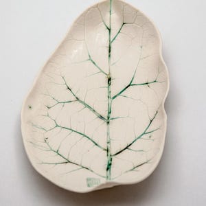 Puede incluir: Un plato de cerámica con forma de hoja en color blanco roto que presenta un patrón de venas de color verde oscuro que se ramifica desde el centro. La superficie tiene una textura de esmalte craquelado y una pequeña marca verde en el borde.