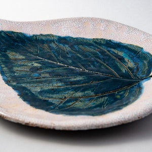 May include: A ceramic leaf-shaped dish with a textured, off-white exterior and a deep blue leaf design in the centre. The leaf's veins are visible, and the dish has a crackled finish, adding to its rustic charm.