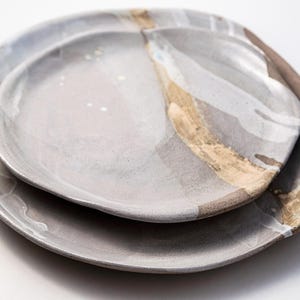 May include: Two ceramic plates, one nested inside the other. The plates are primarily gray with a marbled effect, accented by streaks of tan and white. The edges have a rough, organic shape, suggesting a handmade quality.