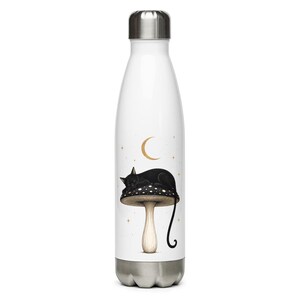 Puede incluir: Una botella de agua de acero inoxidable blanca con tapa y base plateadas. La botella presenta una ilustración gráfica de un gato negro durmiendo sobre un hongo con manchas, acompañado de una luna creciente dorada y pequeñas estrellas.