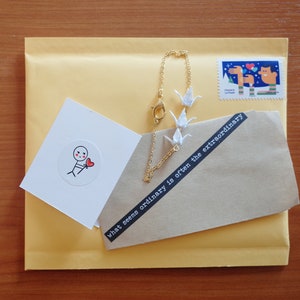 May include: A brown envelope with a white card and a gold chain bracelet with white paper cranes. The card has a simple drawing of a stick figure holding a red heart. The envelope has a black and white sticker with the text "what seems ordinary is often the extraordinary".