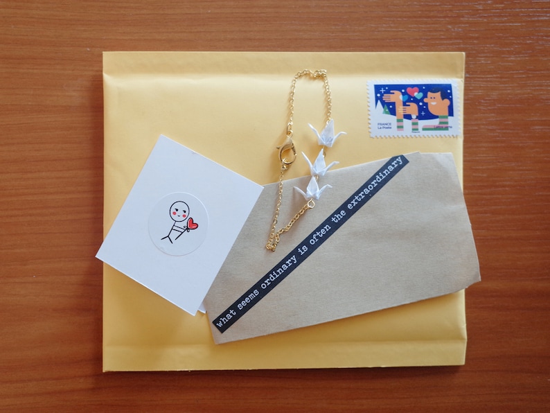 May include: A brown envelope with a white card and a gold chain bracelet with white paper cranes. The card has a simple drawing of a stick figure holding a red heart. The envelope has a black and white sticker with the text "what seems ordinary is often the extraordinary".