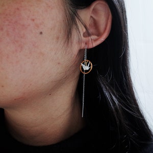 Profile view of a woman with dark hair wearing a black turtleneck and a unique earring featuring a long, delicate chain with a white origami crane encircled in a golden hoop.