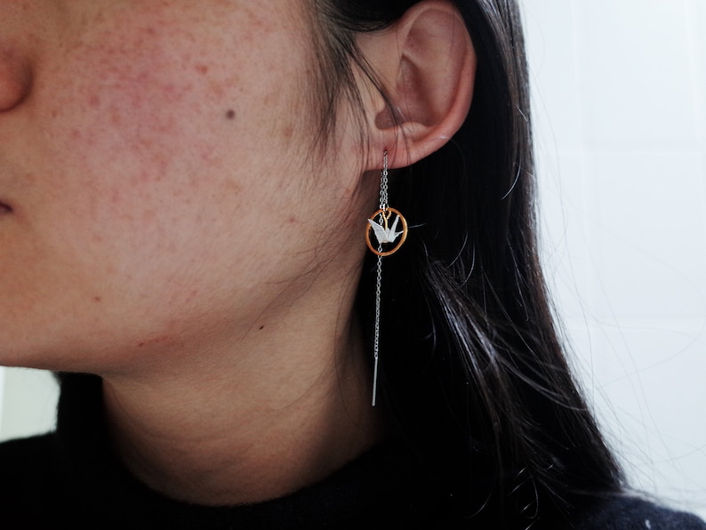 Profile view of a woman with dark hair wearing a black turtleneck and a unique earring featuring a long, delicate chain with a white origami crane encircled in a golden hoop.