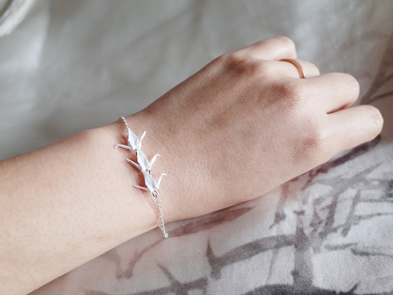 May include: A silver chain bracelet with three white origami cranes attached to it.