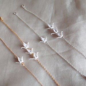 May include: Three delicate gold and silver chain bracelets with origami paper cranes hanging from them. The bracelets are on a white fabric background.
