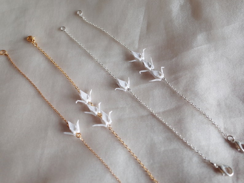 May include: Three delicate gold and silver chain bracelets with origami paper cranes hanging from them. The bracelets are on a white fabric background.