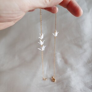 May include: Two gold chains with white origami cranes hanging from them. The chains are on a white fabric background.