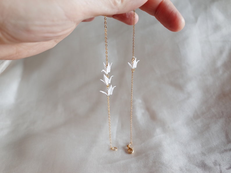 May include: Two gold chains with white origami cranes hanging from them. The chains are on a white fabric background.