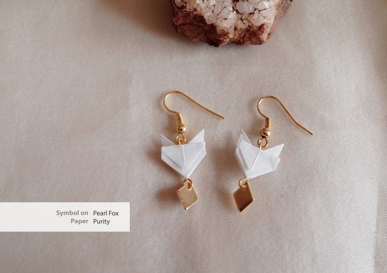 Pu&ograve; includere: Un paio di orecchini dorati con ciondoli a forma di volpe origami bianca e dettagli quadrati dorati. Gli orecchini sono su uno sfondo beige. Simbolo su Pearl Fox Paper Purity