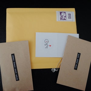 The image displays thoughtful product packaging, including a yellow mailing envelope with a stamp, kraft envelopes with motivational phrases, and a charming card featuring a simple stick figure with a heart.