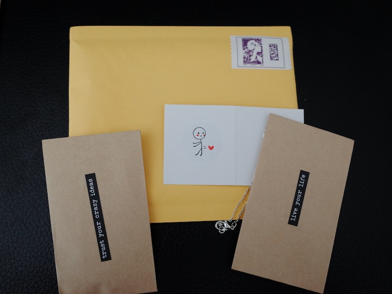 The image displays thoughtful product packaging, including a yellow mailing envelope with a stamp, kraft envelopes with motivational phrases, and a charming card featuring a simple stick figure with a heart.
