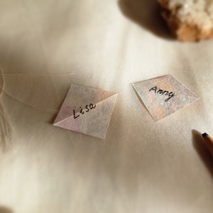 Two small washi paper notes with "Lisa" and "Anny" handwritten on them, resting on a textured surface with a pencil and a stone in soft lighting.