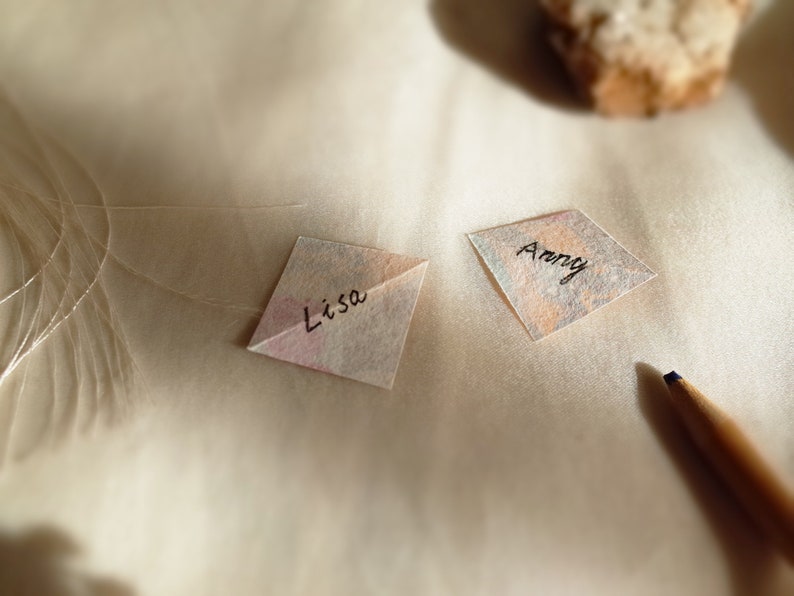 Two small washi paper notes with "Lisa" and "Anny" handwritten on them, resting on a textured surface with a pencil and a stone in soft lighting.