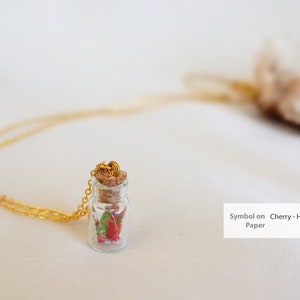 A delicate necklace with a tiny glass bottle pendant containing a red and green origami crane, symbolizing hope, capped with a cork and attached to a golden chain, intended as a gift for new mothers.