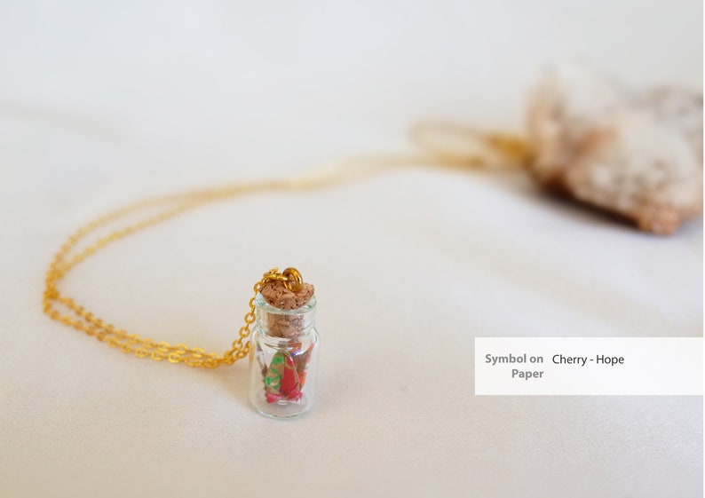 A delicate necklace with a tiny glass bottle pendant containing a red and green origami crane, symbolizing hope, capped with a cork and attached to a golden chain, intended as a gift for new mothers.