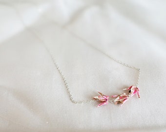 Peut inclure: Un collier en chaîne argentée avec trois grues en origami roses suspendues. Le collier est sur un fond de tissu blanc.