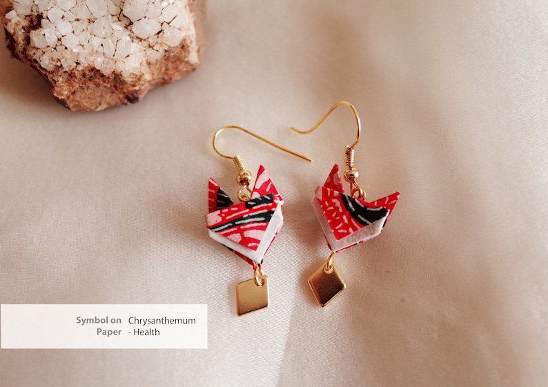 Pu&ograve; includere: Un paio di orecchini dorati con ciondoli a forma di volpe origami rosso, bianco e nero. I ciondoli sono decorati con un motivo di crisantemo. Gli orecchini hanno un pendente quadrato dorato.