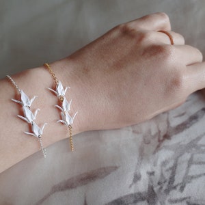 A wrist with two bracelets: a gold chain with three origami crane charms and a silver chain with three origami crane charms, on a neutral background.