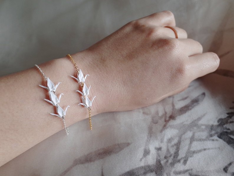 A wrist with two bracelets: a gold chain with three origami crane charms and a silver chain with three origami crane charms, on a neutral background.