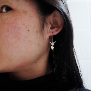 A woman is shown in profile wearing a black turtleneck, with a long, elegant gold chain earring featuring a small origami crane that delicately complements her minimalist style.
