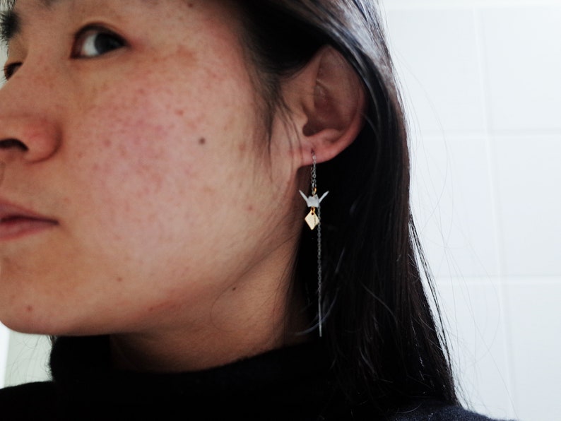 A woman is shown in profile wearing a black turtleneck, with a long, elegant gold chain earring featuring a small origami crane that delicately complements her minimalist style.