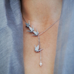 Könnte beinhalten: Eine silberne Halskette mit einer zarten Kette und drei Origami-Kranich-Anhängern. Die Halskette hat einen einzelnen Perlenanhänger am Ende.