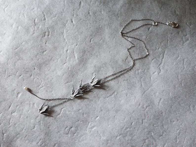Może przedstawiać: Srebrny naszyjnik z delikatnym łańcuszkiem i zawieszką przedstawiającą kilka ptak&oacute;w origami. Naszyjnik ma perłowy akcent na końcu.