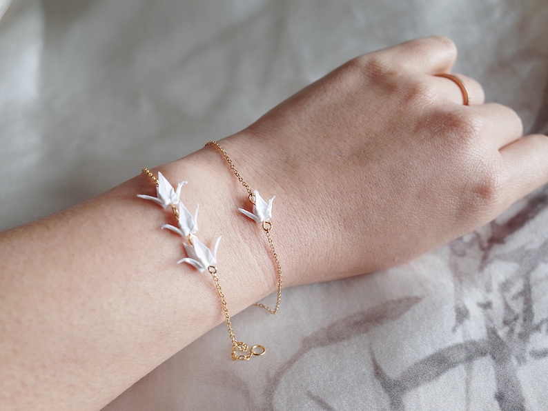 A slender wrist adorned with two delicate bracelets: one featuring a single crane charm, the other with three crane charms, both with a gold chain.