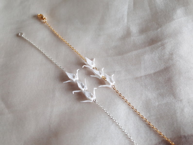 May include: Two delicate bracelets with origami paper crane charms. One bracelet has a silver chain and the other has a gold chain.