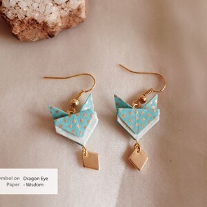 Pu&ograve; includere: Un paio di orecchini origami con un design a motivi blu e bianchi e dettagli dorati. Gli orecchini hanno la forma di un simbolo di occhio di drago e hanno un piccolo ciondolo a forma di diamante dorato che pende dalla parte inferiore.
