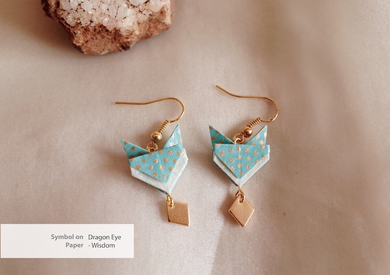 Pu&ograve; includere: Un paio di orecchini origami con un design a motivi blu e bianchi e dettagli dorati. Gli orecchini hanno la forma di un simbolo di occhio di drago e hanno un piccolo ciondolo a forma di diamante dorato che pende dalla parte inferiore.