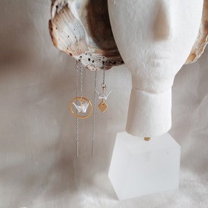 A mannequin displays asymmetrical earrings: one with a white origami crane within a gold circle, the other a diamond shape, both dangling from silver chains against a soft, textured backdrop.