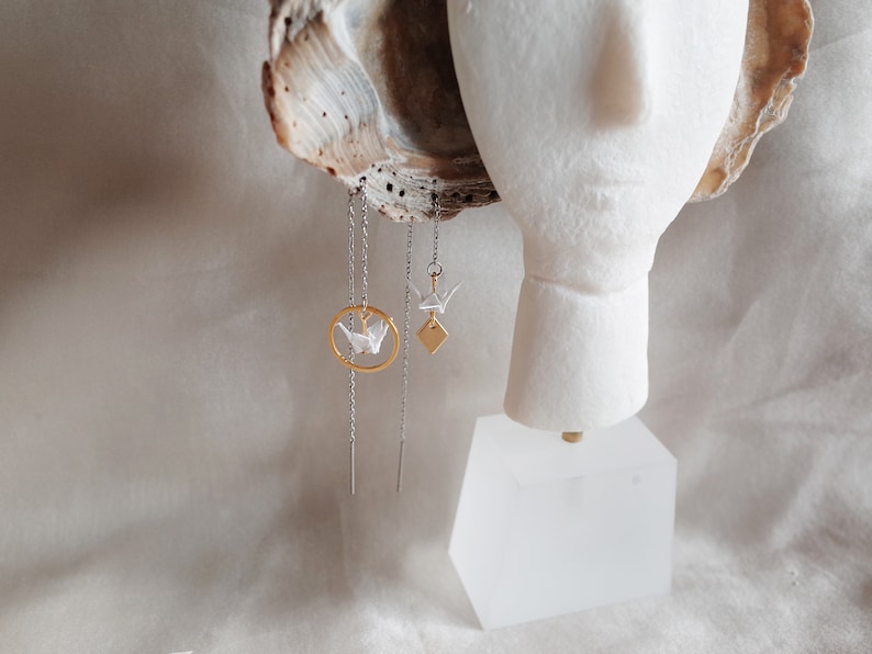 A mannequin displays asymmetrical earrings: one with a white origami crane within a gold circle, the other a diamond shape, both dangling from silver chains against a soft, textured backdrop.