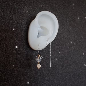 Può includere: Un paio di orecchini a catena d'argento con un piccolo ciondolo a forma di gru origami e un ciondolo a barra sottile d'argento appeso alle catene.