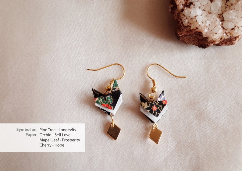 Pu&ograve; includere: Un paio di orecchini pendenti dorati con fiori di carta origami in nero, bianco e verde. I fiori sono decorati con perline dorate e gli orecchini hanno un ciondolo quadrato in basso.