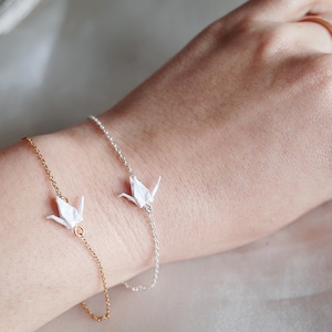 A wrist with two bracelets: a gold chain with one origami crane charm and a silver chain with one origami crane charm, on a neutral background.