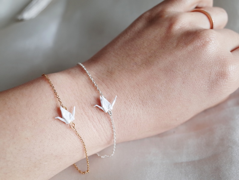 A wrist with two bracelets: a gold chain with one origami crane charm and a silver chain with one origami crane charm, on a neutral background.
