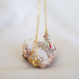 A necklace with a miniature glass bottle containing a tiny red and green origami crane, suspended on a golden chain, delicately resting against a cluster of sparkling white crystals.